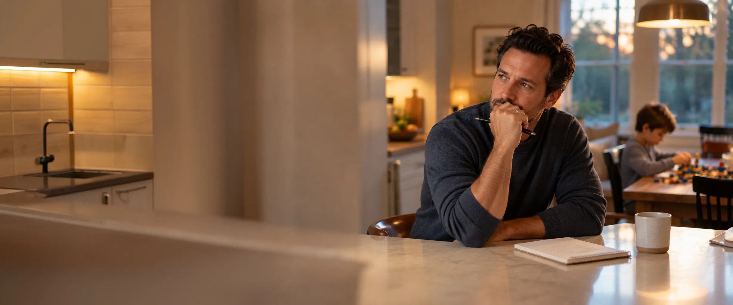A father sits at the kitchen counter in the evening, thinking, with a notepad in front of him, while his son builds at the table behind him.