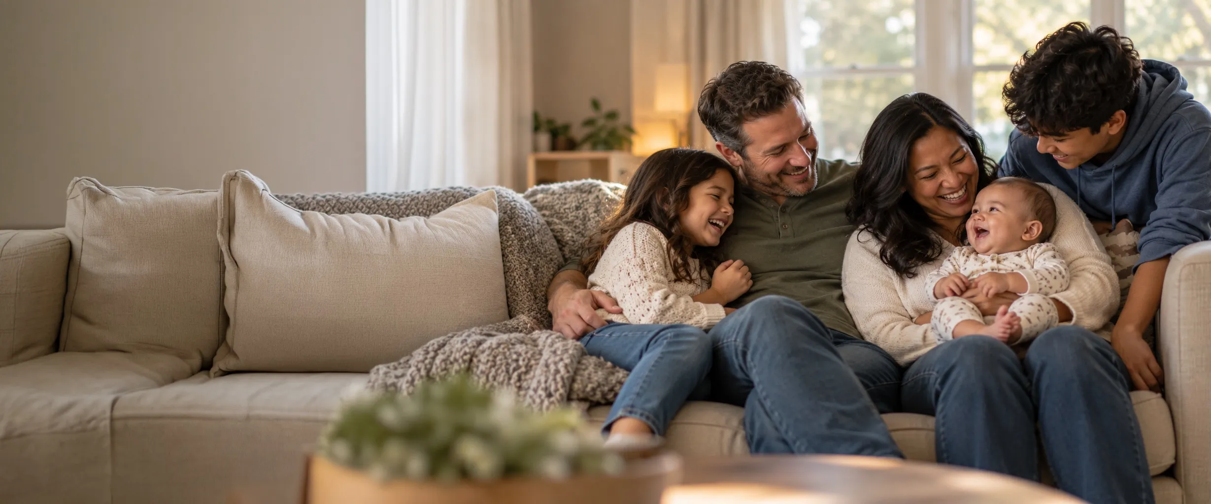 A family with a baby, a young daughter, and a teenage son laughs together on a living-room couch.